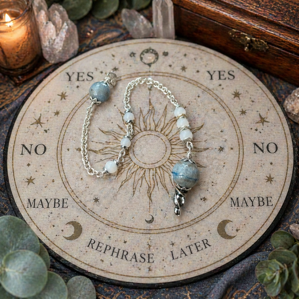 Jena Jewelry pendulum with blue aventurine, snow jade, and hematite beads on a chainmaille handle, resting on a divination board.