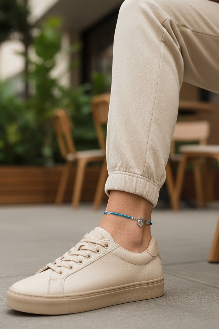 Jena Jewelry handmade blue seed bead anklet with a silver heart charm, worn with beige sneakers.