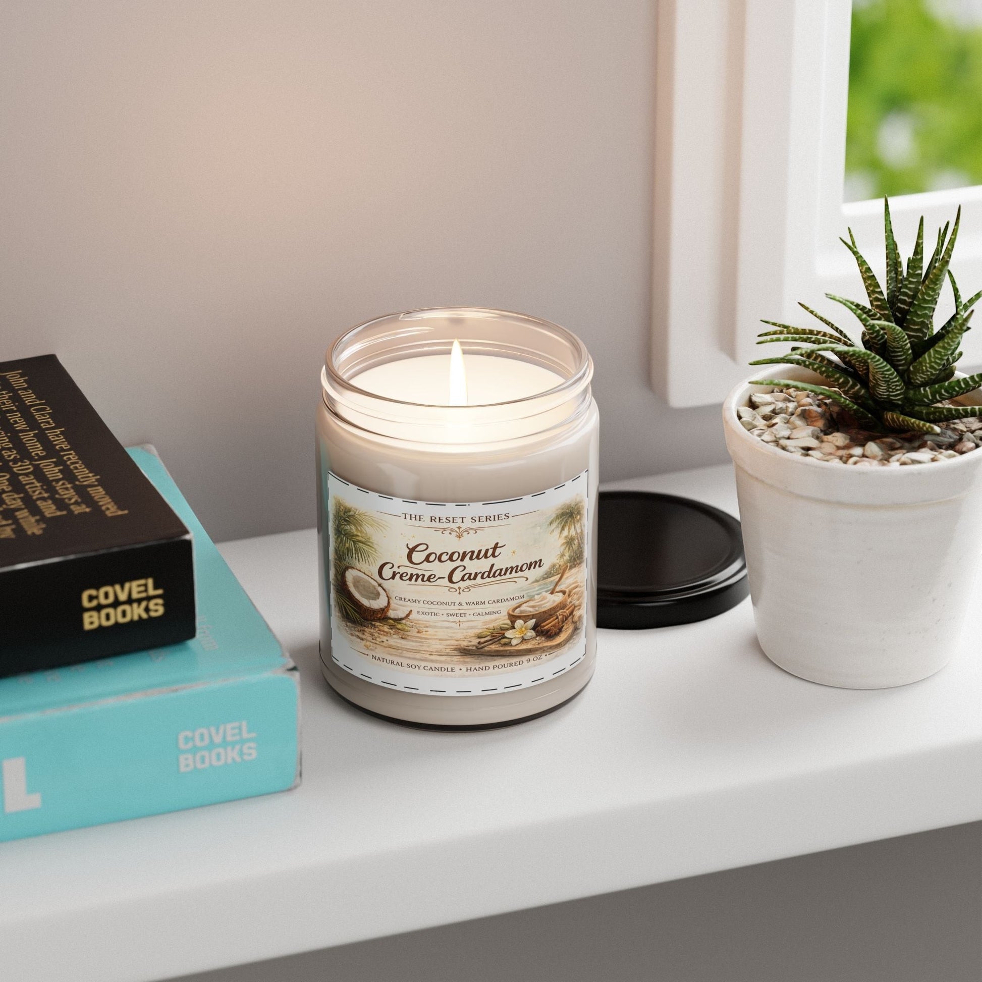 Jena Jewelry Coconut Cardamom soy candle in a glass jar with a lit wick, next to books and a potted plant.