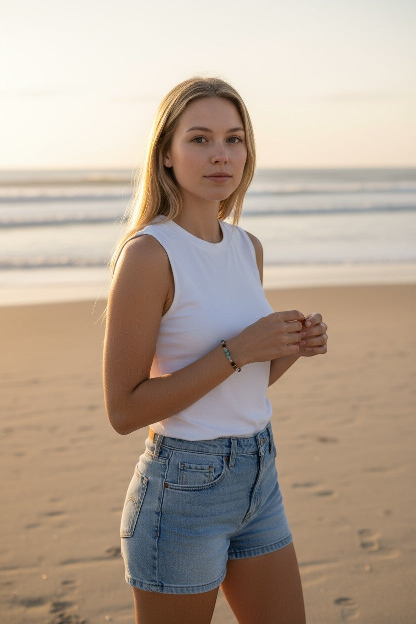 Woman wearing a white tank top and denim shorts on a beach, with a multi-stone bracelet on her wrist. Jena Jewelry.