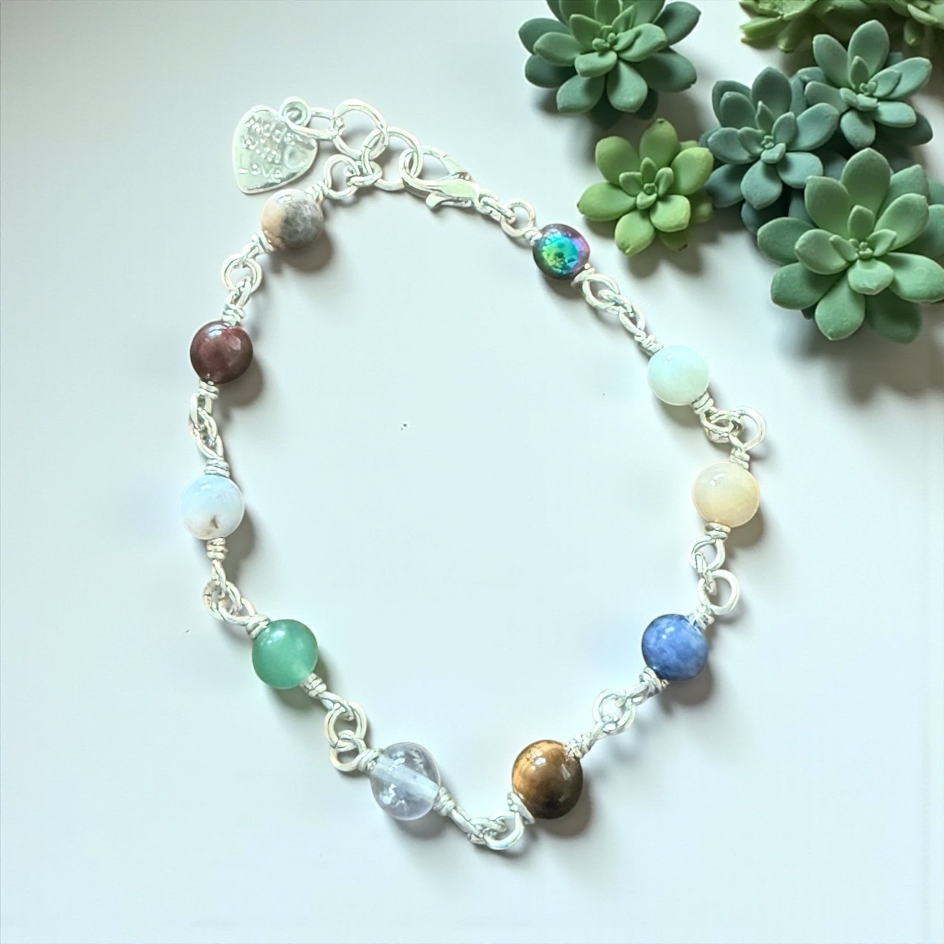 Jena Jewelry multi-stone bracelet with silver wire wrapping and a heart charm. Features various colorful stones including green, blue, brown, and iridescent.