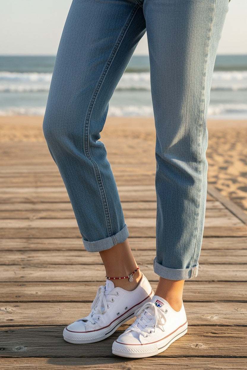 Jena Jewelry handmade red and white seed bead anklet with a silver charm, worn with jeans and sneakers on a boardwalk.