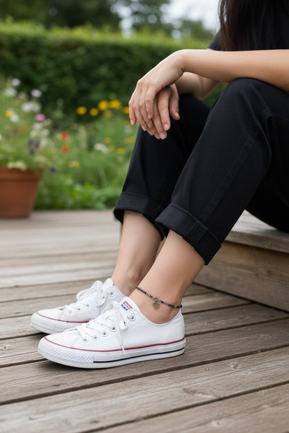 Jena Jewelry handmade seed bead anklet with a heart charm, featuring purple and gold beads, worn on a woman's ankle with black pants and white sneakers.