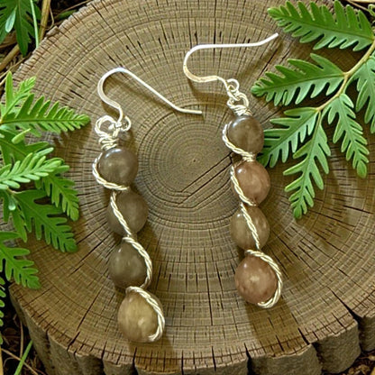 Jena Jewelry stacked drop earrings with brown and tan polished stones, wire-wrapped on silver-tone hooks. Natural wood and fern backdrop.