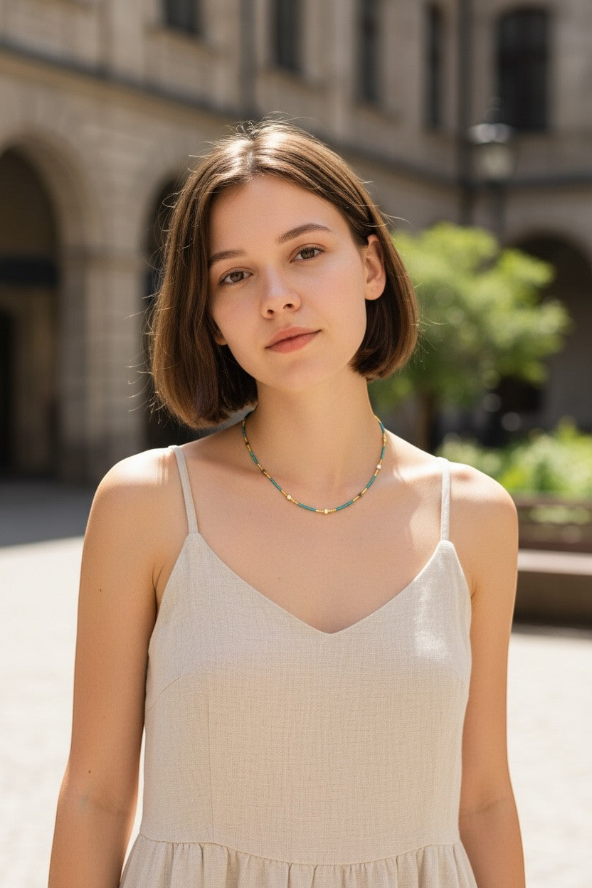 Jena Jewelry teal and gold seed bead necklace on a young woman wearing a beige linen dress outdoors.