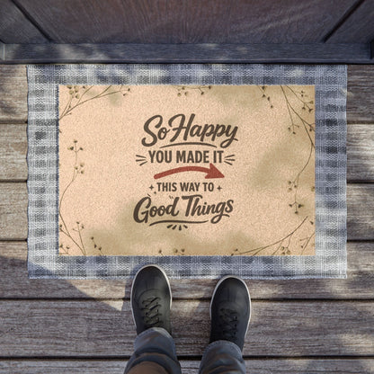 Jena Jewelry "So Happy You Made It" doormat with a beige coir surface, dark text, and a blue checkered border. Features botanical accents.