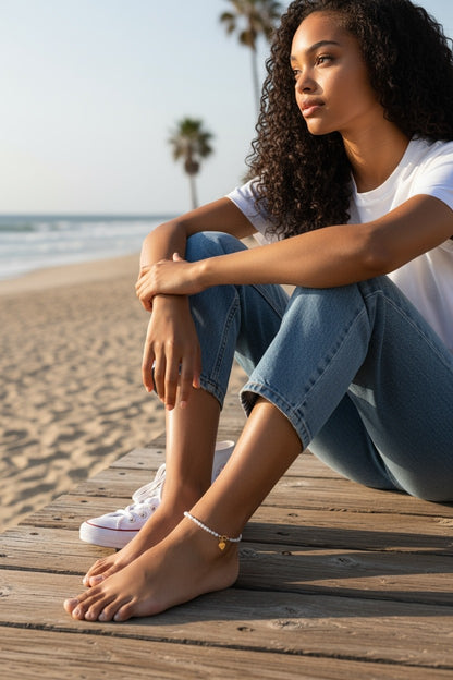 Jena Jewelry white seed bead anklet with gold heart charm on a woman's ankle at the beach.