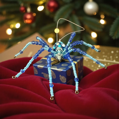 Jena Jewelry blue beaded spider ornament with silver accents, sitting on a gift box with a Christmas tree in the background.