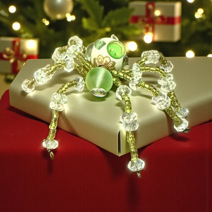 Jena Jewelry handcrafted spider ornament with green bead body and clear crystal legs, on a gift box with Christmas tree background.
