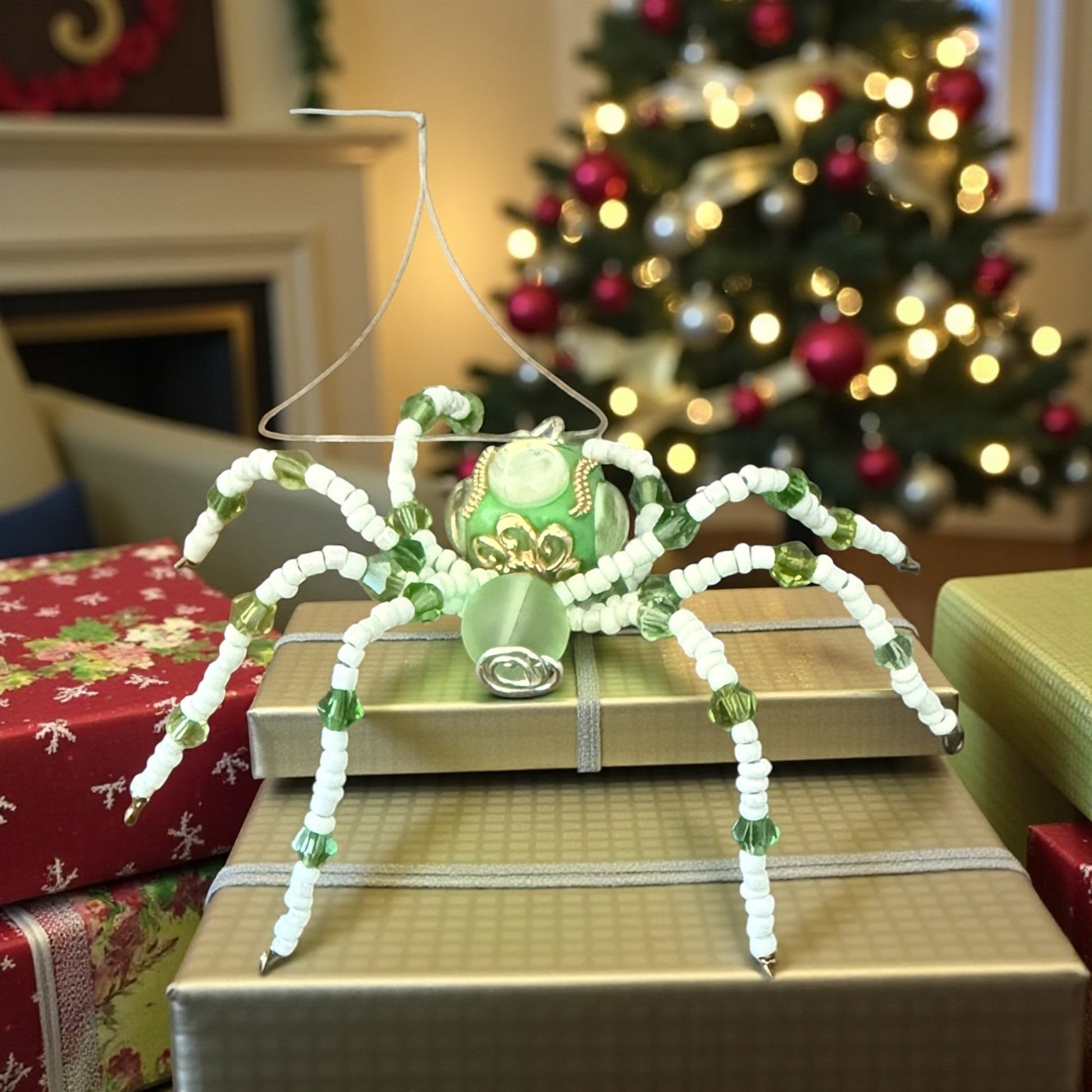 Jena Jewelry green and white beaded spider ornament with a glass body, displayed on gift boxes with a Christmas tree in the background.