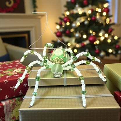 Jena Jewelry green and white beaded spider ornament with a glass body, displayed on gift boxes with a Christmas tree in the background.