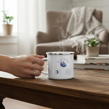 White enamel camping mug with "Trust the Pull" text and moon phase graphics. A hand holds the mug, which is steaming.