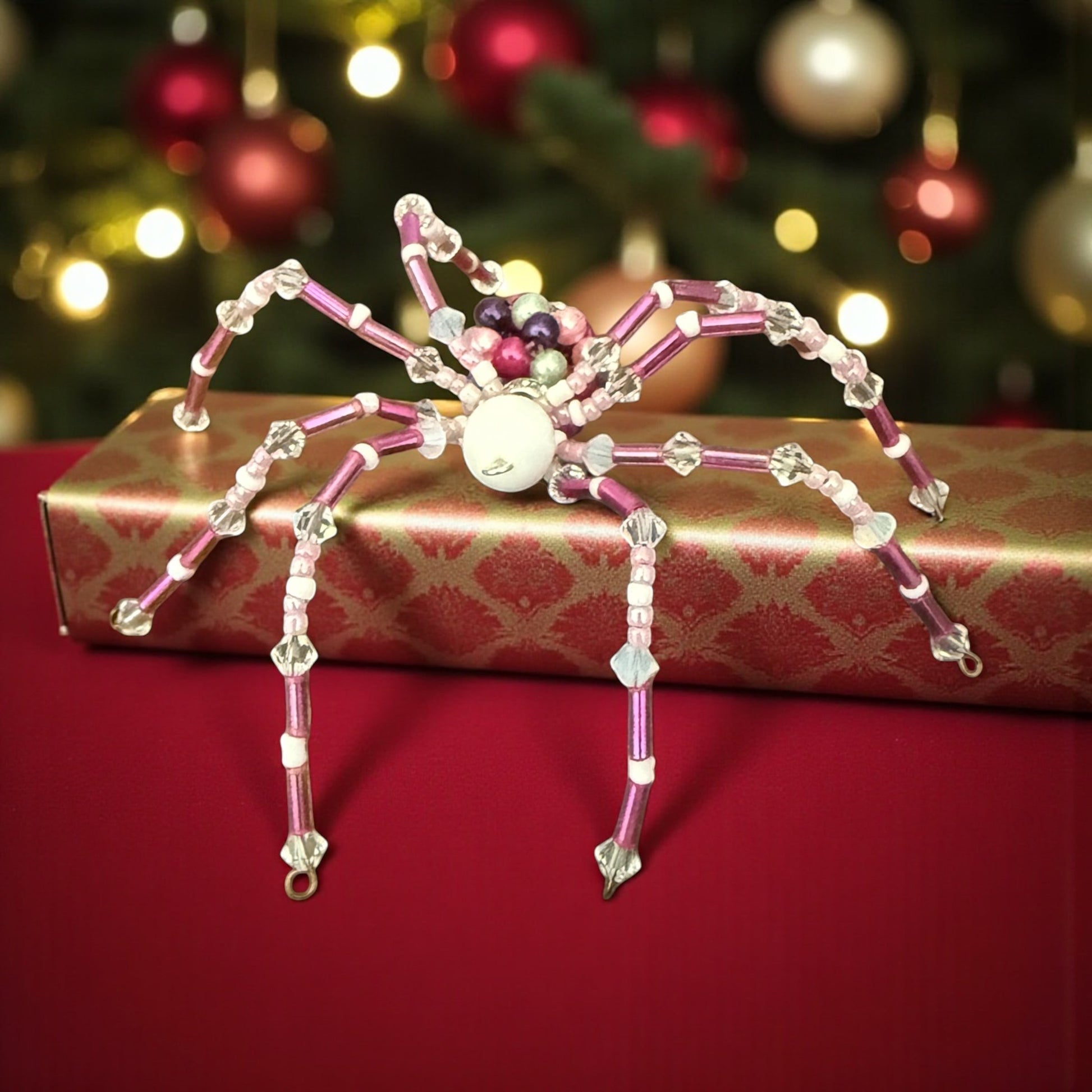 Jena Jewelry handcrafted pink and clear beaded spider ornament with a white body, sitting on a gold and red patterned gift box.