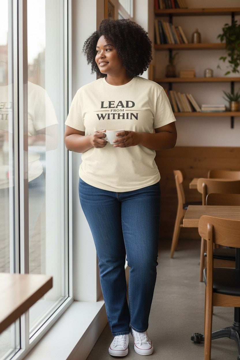 Woman wearing neutral beige “Lead From Within” t-shirt near a window, a calm and confident everyday look.