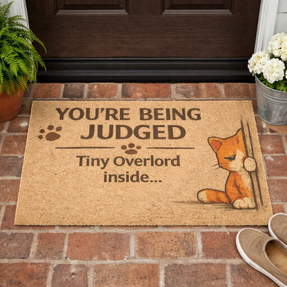 Funny “You’re Being Judged – Tiny Overlord Inside” cat doormat displayed at a front door, perfect for cat lovers with a sense of humor.